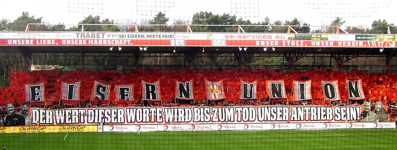 The packed Waldseite terrace at Union Berlin's Alte Försterei, holding up a red and white tifo reading Eisern Union